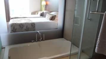 Um banheiro moderno com chuveiro em box de vidro, uma banheira livre e um quarto visível com cama e mesa de cabeceira.
