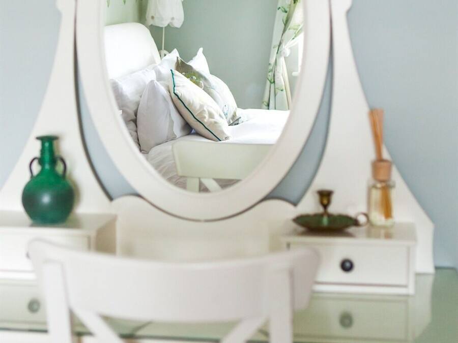A white vanity with a round mirror, a green vase, and a small table with a candle and a decorative item.