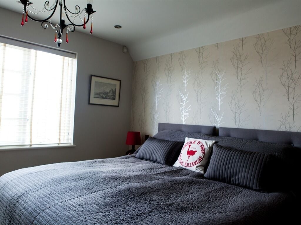 Un dormitorio con una cama grande, una araña de cristal, una ventana con persianas y un papel pintado con motivos de árboles.