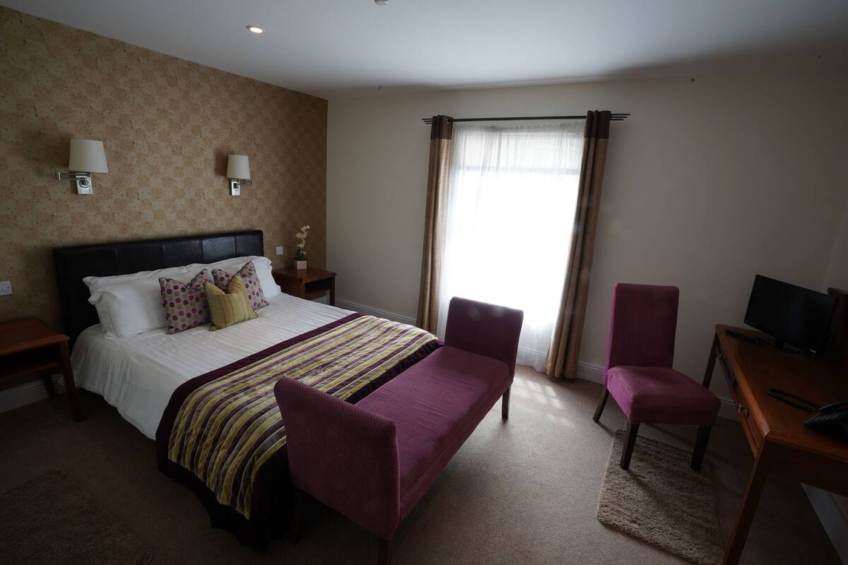 A hotel room with a bed, two chairs, a desk with a TV, and a window with curtains.