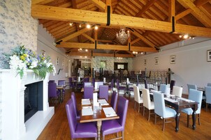 Restaurante com vigas de madeira, cadeiras roxas e brancas, e uma lareira.