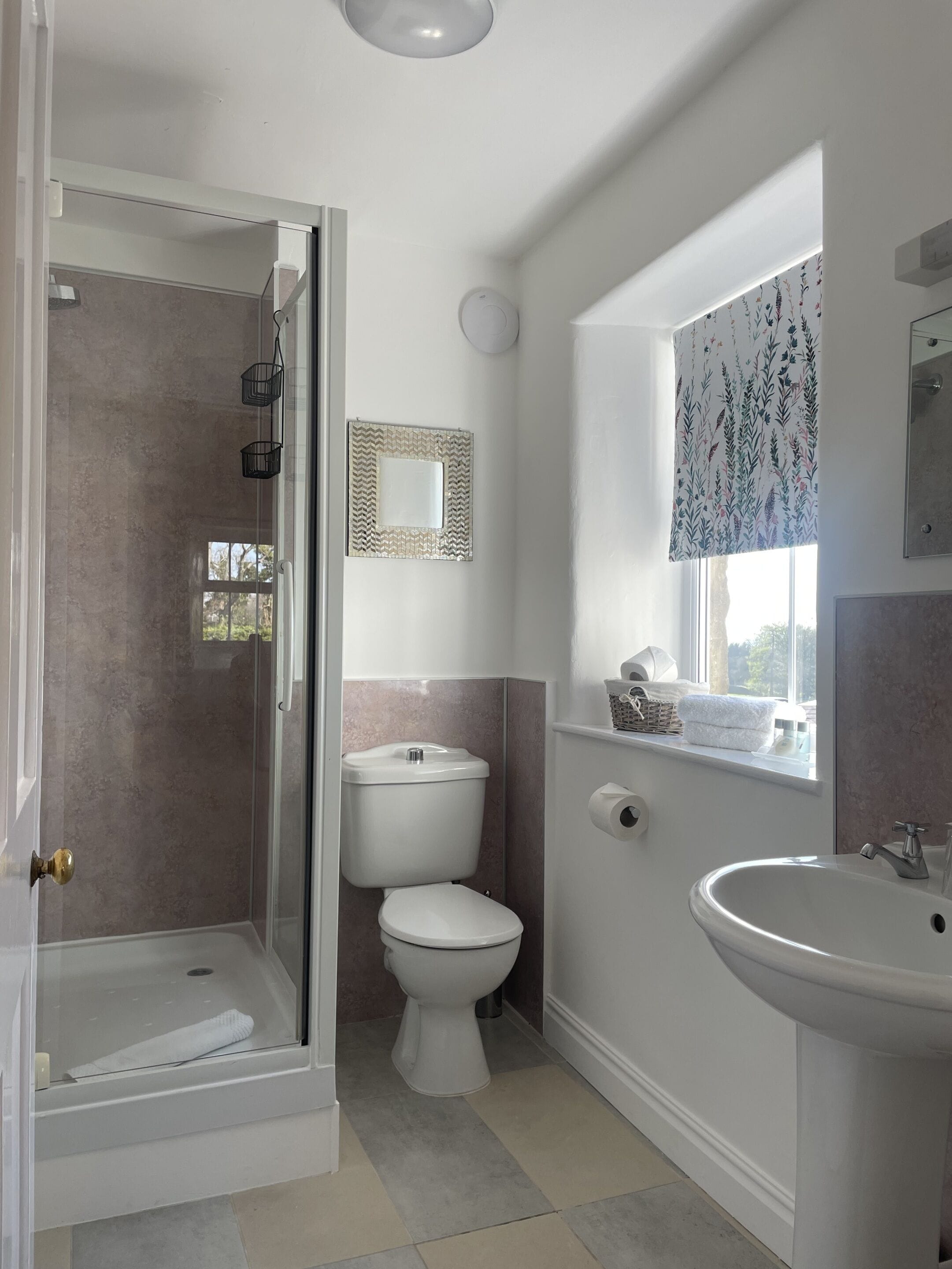Un baño con ducha de vidrio, inodoro, lavamanos y ventana con cortina de motivos florales.