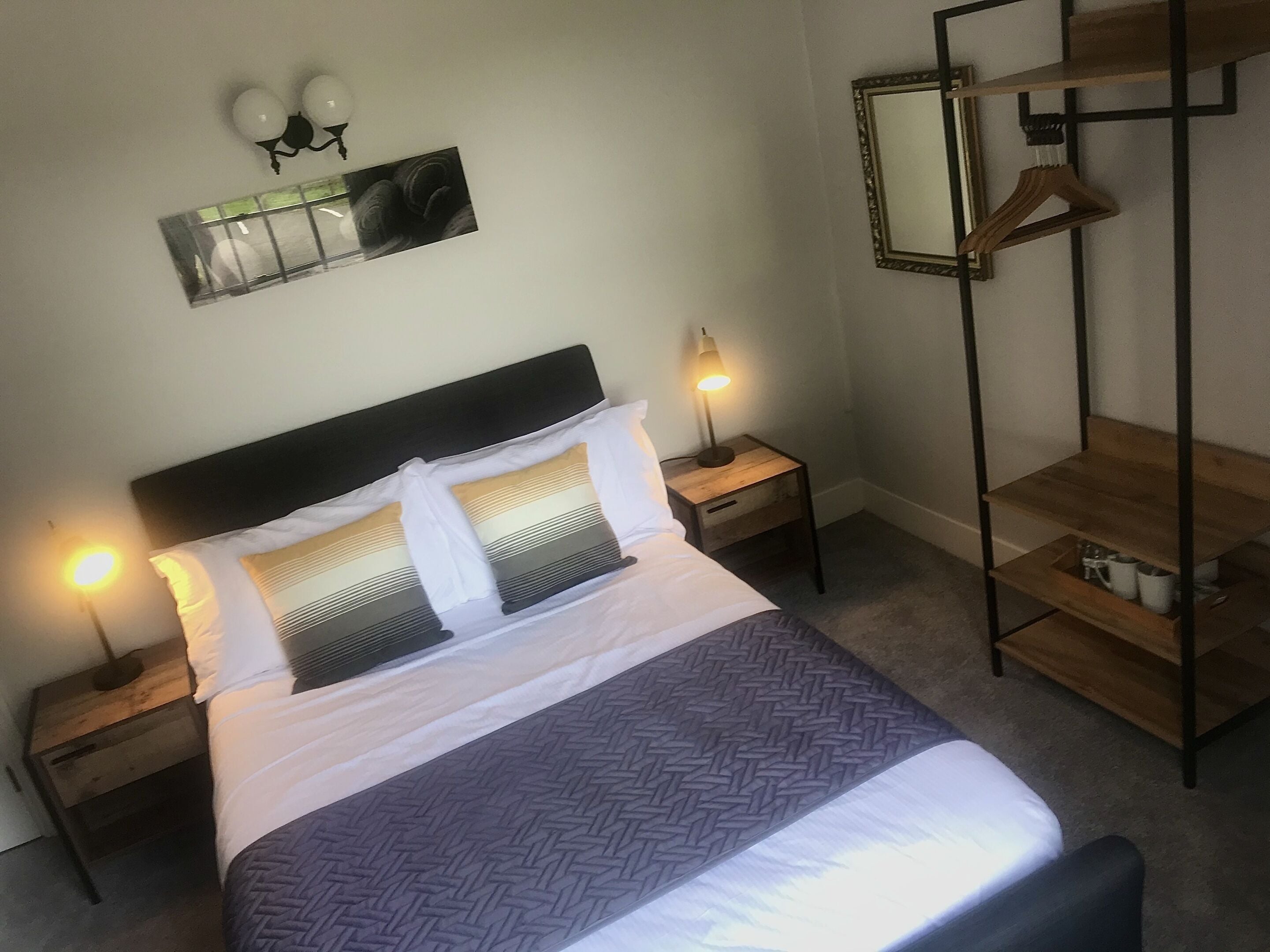 Una cama bien hecha con cabecera, dos mesitas de noche con lámparas, una mesita de noche de madera, un espejo y un perchero de madera.