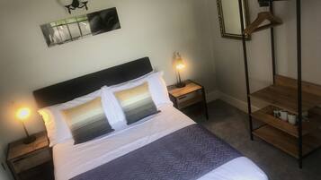 Una cama bien hecha con cabecera, dos mesitas de noche con lámparas, una mesita de noche de madera, un espejo y un perchero de madera.