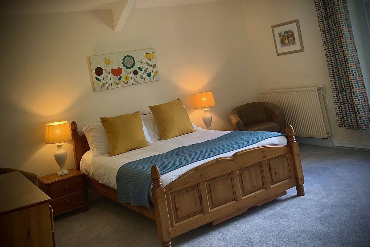 A bedroom with a wooden bed, two bedside lamps, a chair, a radiator, and a wall art piece.