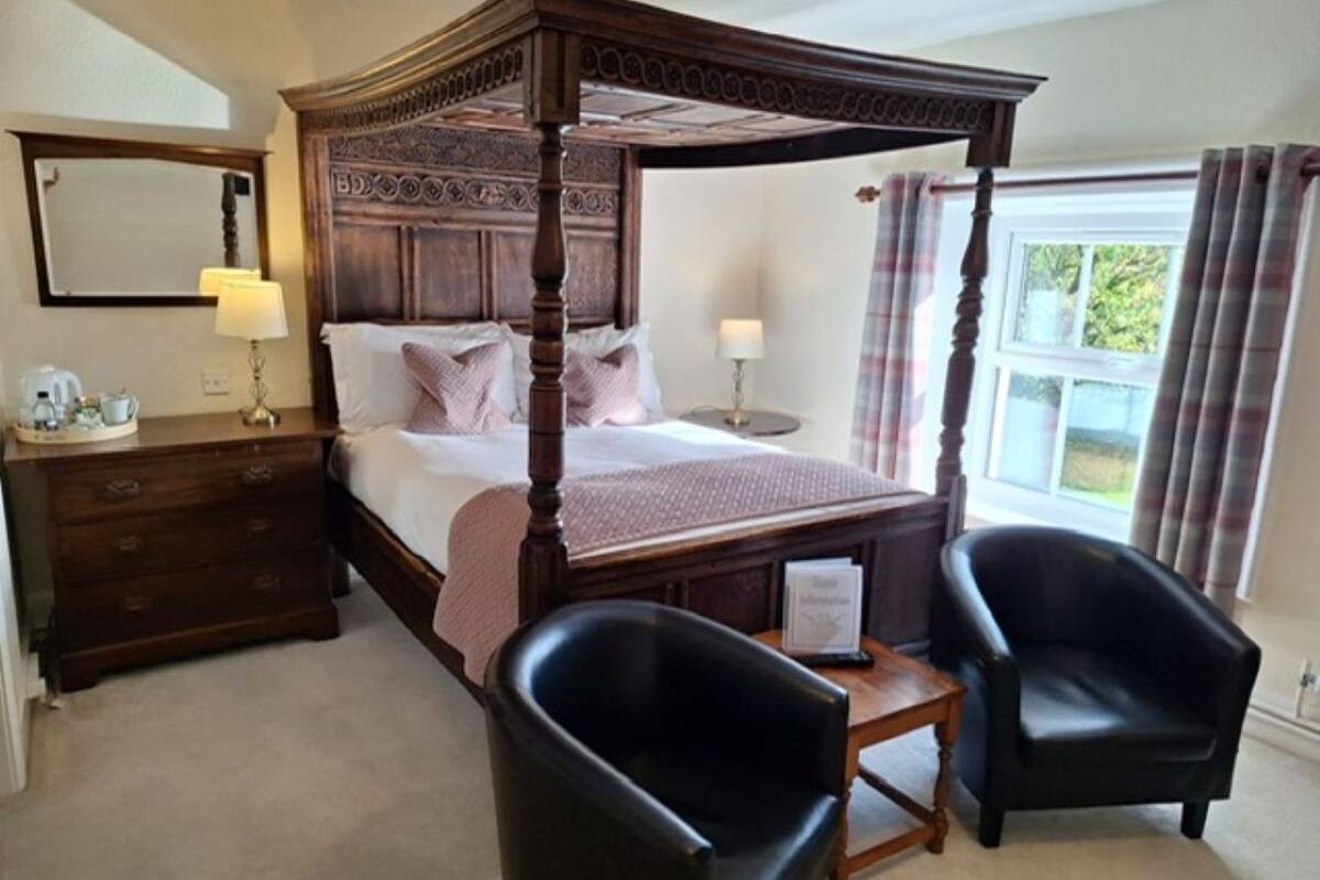 A four-poster bed with a wooden headboard, two bedside tables, a nightstand, and two black leather chairs.