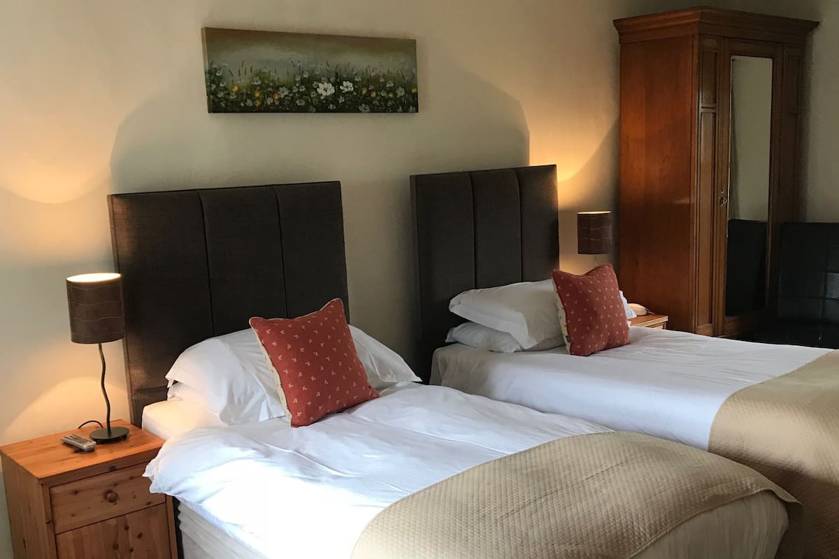 A hotel room with two beds, a nightstand with a lamp, a wooden wardrobe, and a painting on the wall.