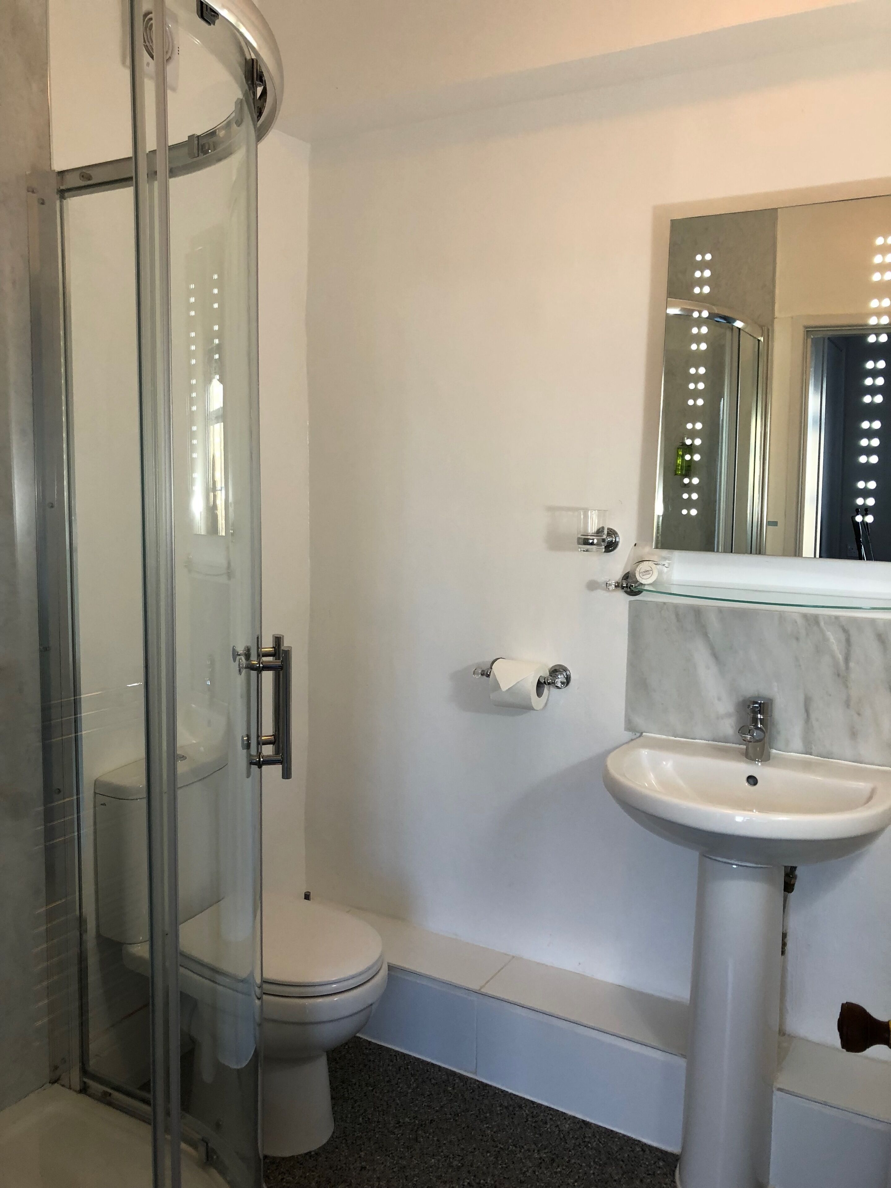 A modern bathroom with a glass shower enclosure, a white toilet, a marble sink, and a mirror with lights.