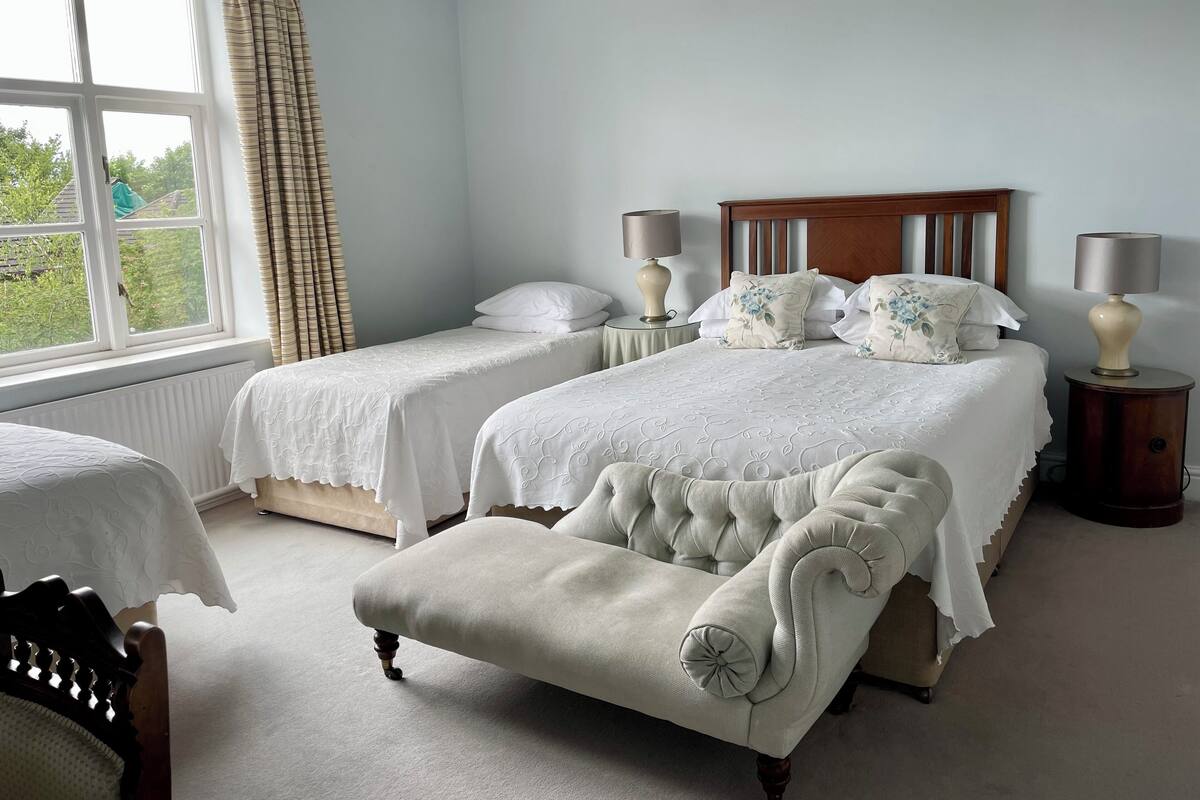 A bedroom with two single beds, a large bed, a wooden headboard, two bedside tables with lamps, and a window with curtains.