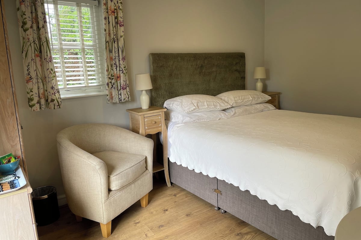 A bedroom with a bed, a nightstand, a chair, a window with curtains, and a wooden floor.