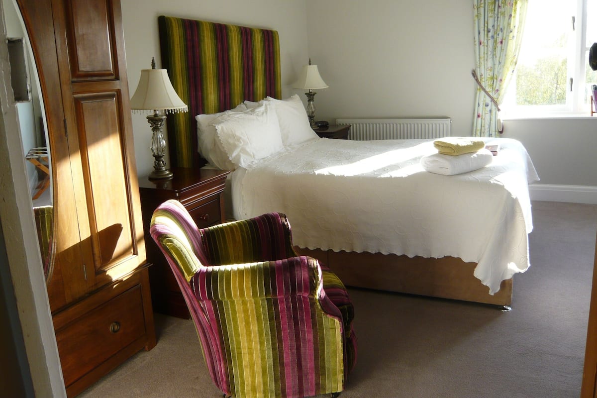 A bedroom with a bed, two bedside lamps, a chair, a wooden wardrobe, and a window with curtains.