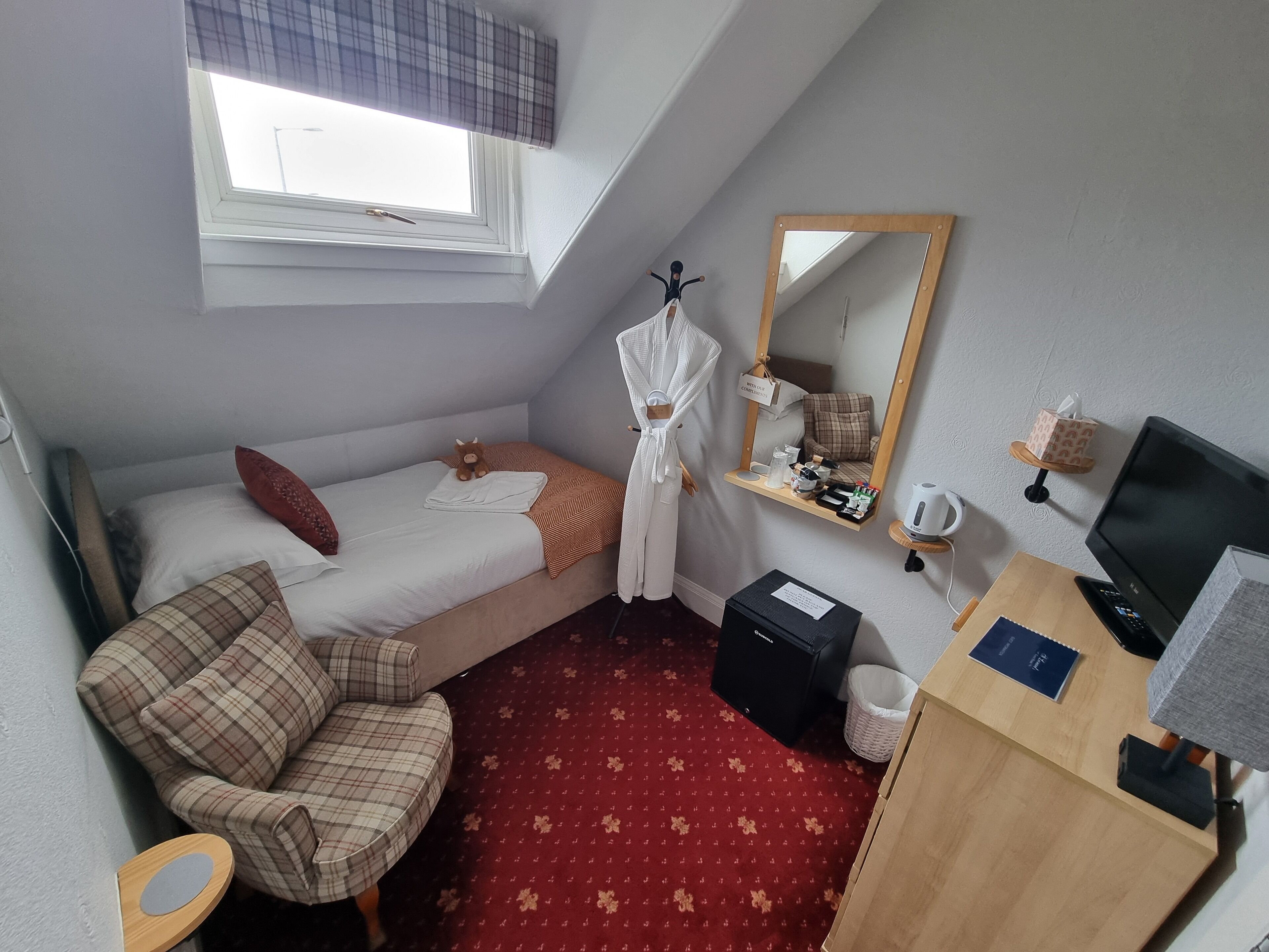 A compact hotel room with a single bed, a small sofa, a desk with a computer, a mirror, and a window with a view.