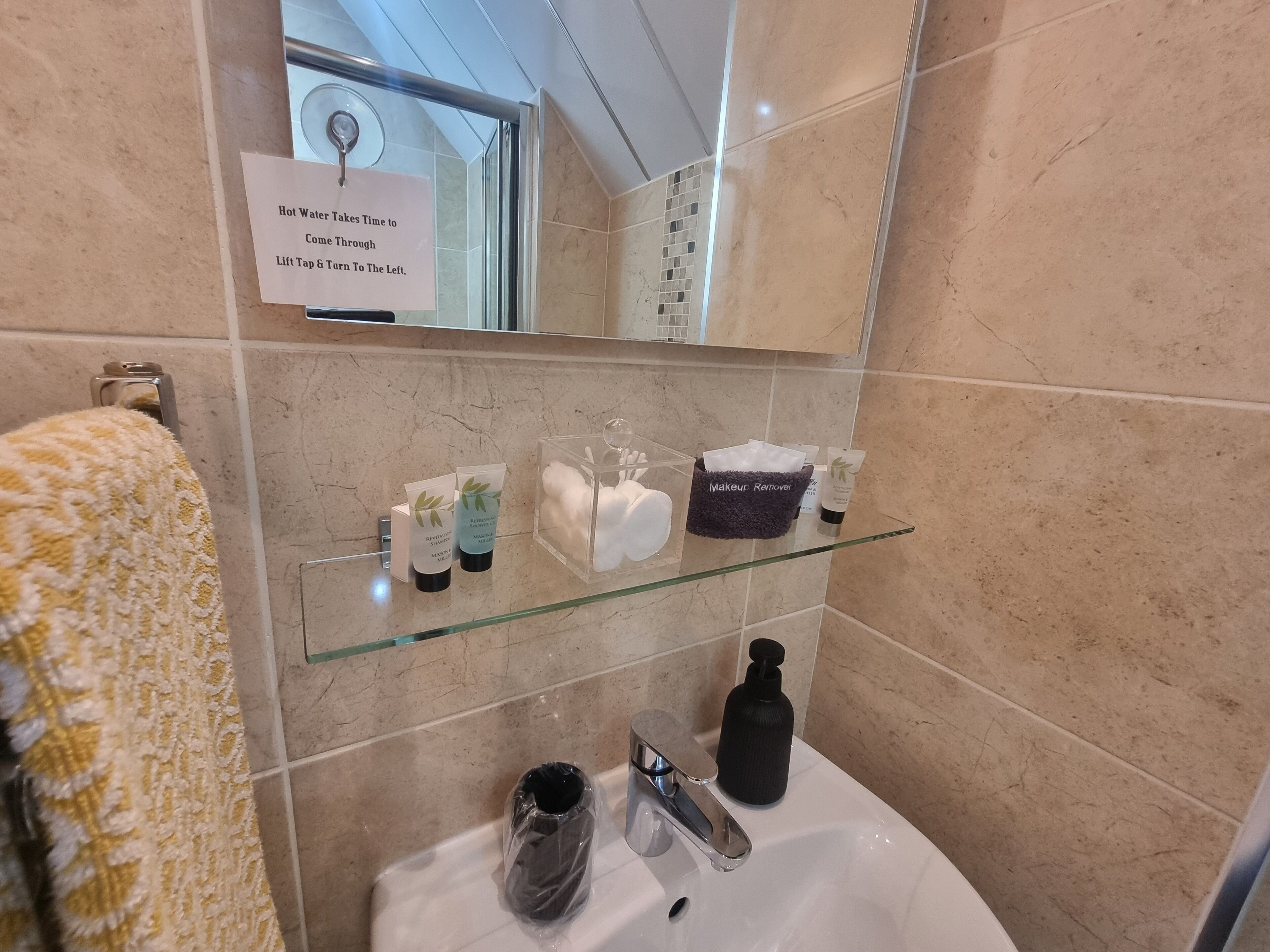 Un baño con un estante de vidrio con artículos de tocador, un perchero con una toalla y un cartel sobre el espejo.