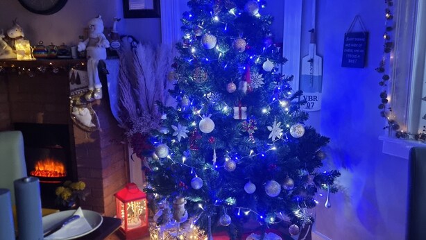 Un sapin de Noël décoré de guirlandes lumineuses et d’ornements, entouré d’un coin salon chaleureux avec une cheminée, une table avec une bougie et une lanterne décorative.