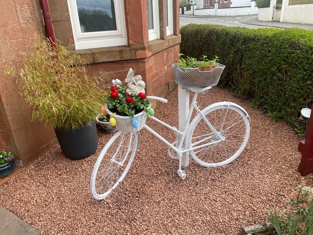 Un vélo blanc, équipé d’un panier et d’une petite plante fixée sur le porte-bagages, est garé sur une surface recouverte de gravier.