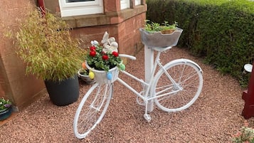 Un vélo blanc, équipé d’un panier et d’une petite plante fixée sur le porte-bagages, est garé sur une surface recouverte de gravier.