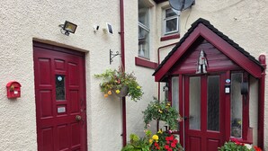 Une porte rouge avec une pancarte indiquant « Entrée des invités », une boîte aux lettres et des plantes en pot devant un bâtiment dont le mur présente une texture particulière et qui est doté d’un petit porche.