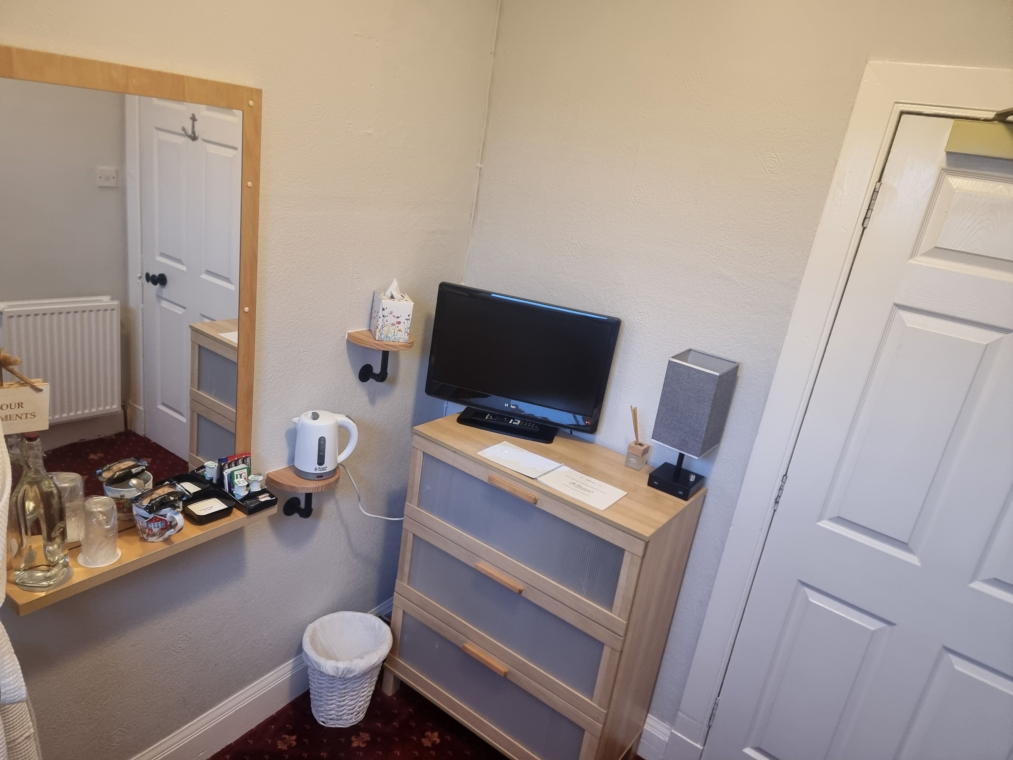 A small, tidy room with a television, a small shelf with a tissue box, a kettle, and a waste bin.