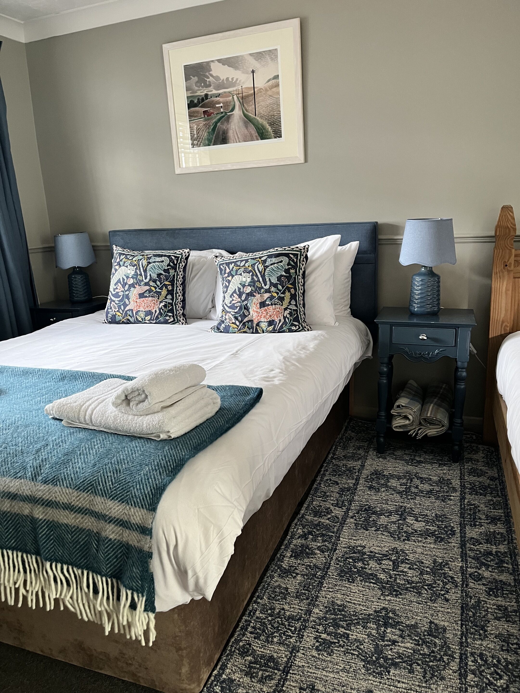 A neatly made bed with white linens and decorative pillows, a blue throw blanket, and a folded towel on the bed. There are two bedside tables with lamps and a framed picture above the bed.
