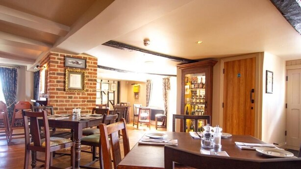 A dining area with wooden tables and chairs, a brick wall with framed pictures, and a wooden door leading to a room with shelves. - The Cherry Tree Rochford (Rochford)