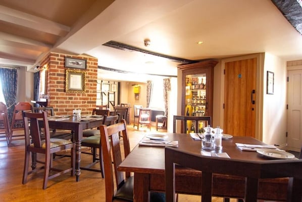 A dining area with wooden tables and chairs, a brick wall with framed pictures, and a wooden door leading to a room with shelves.