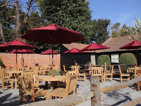 Outdoor wooden tables and chairs with red umbrellas, a brick wall, and a building with a tiled roof.