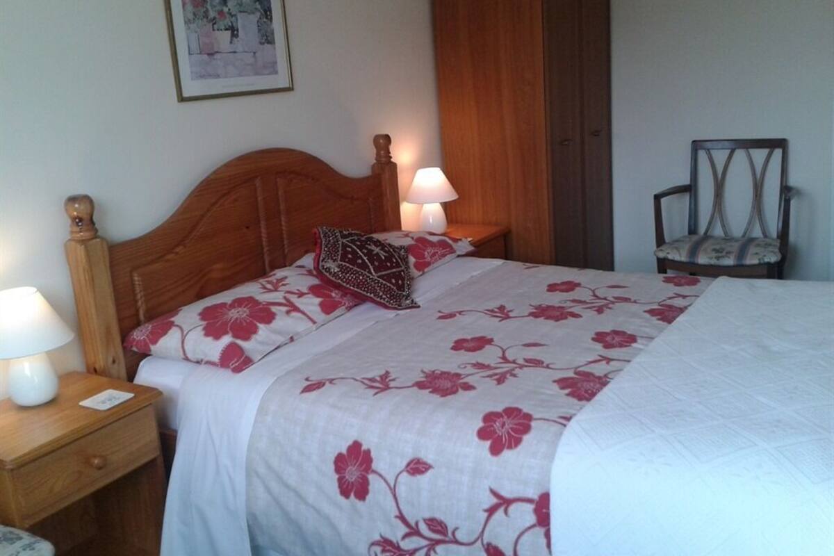 A bedroom with a wooden headboard, a floral bedspread, two bedside lamps, a nightstand, a chair, and a wardrobe.