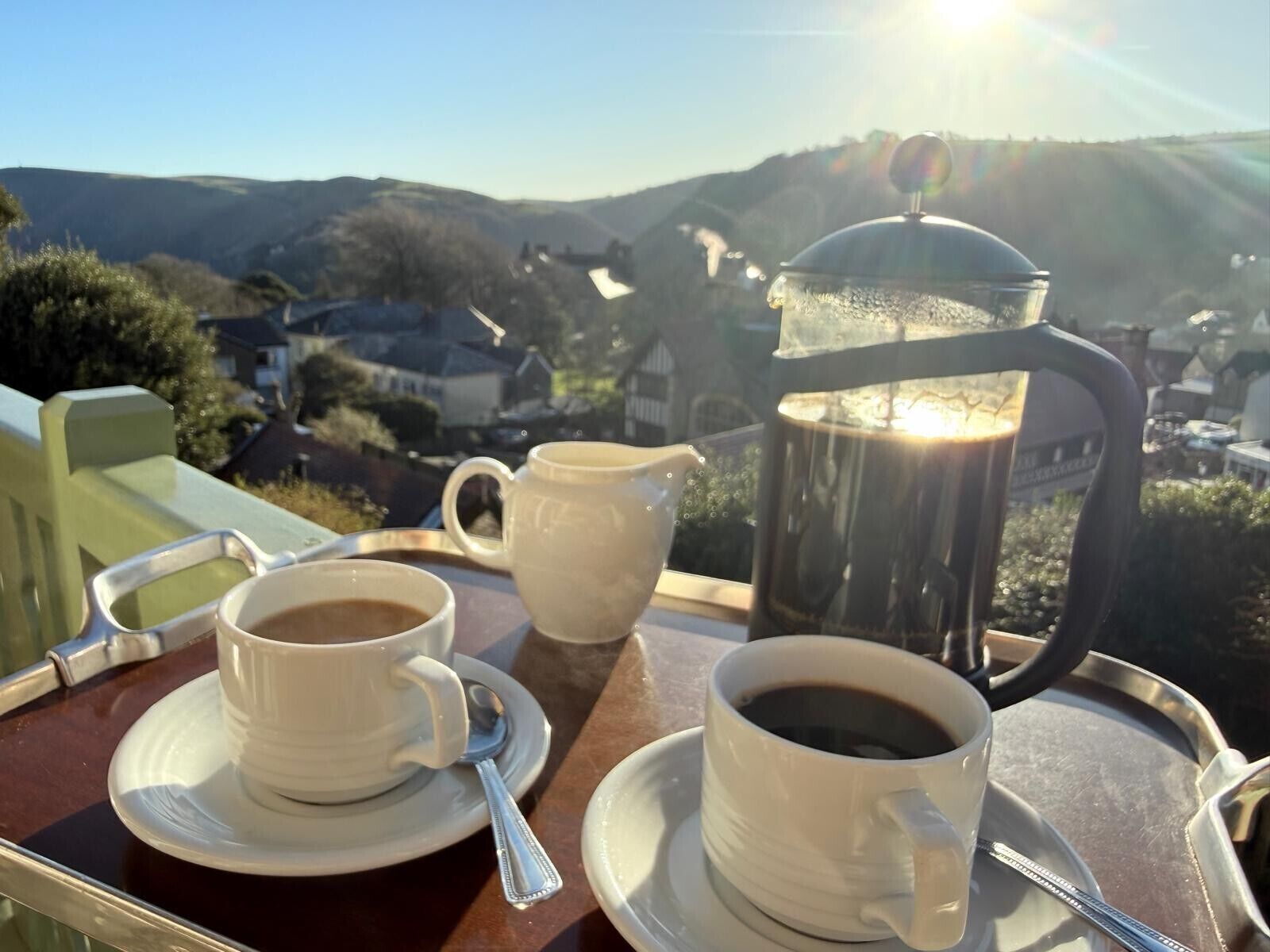 A tray with two cups of coffee and a coffee pot on a table with a view of a village.