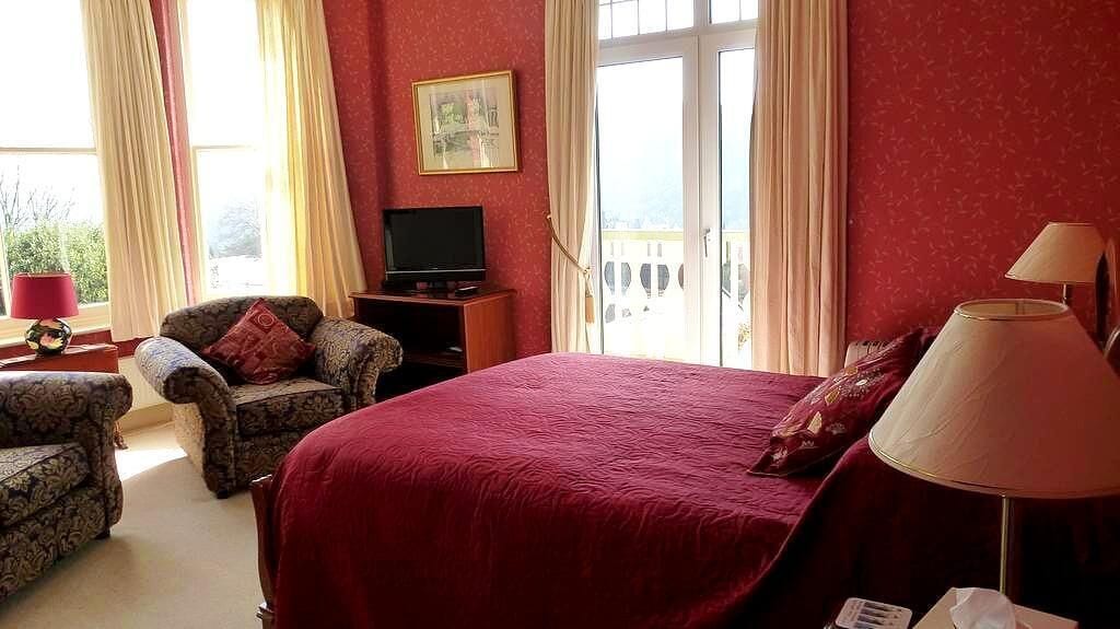 A hotel room with a large bed, a television, a chair, and a balcony with curtains.