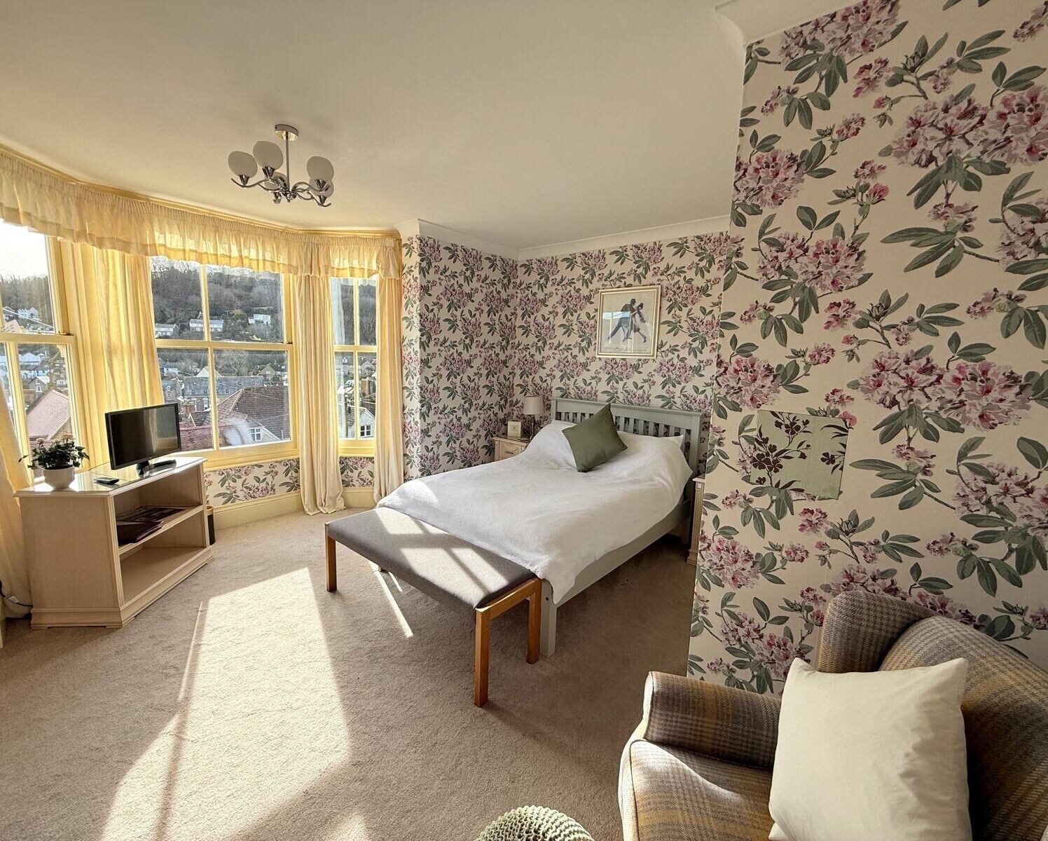 A bedroom with a bed, a TV, a bedside table, a window with curtains, and floral wallpaper.