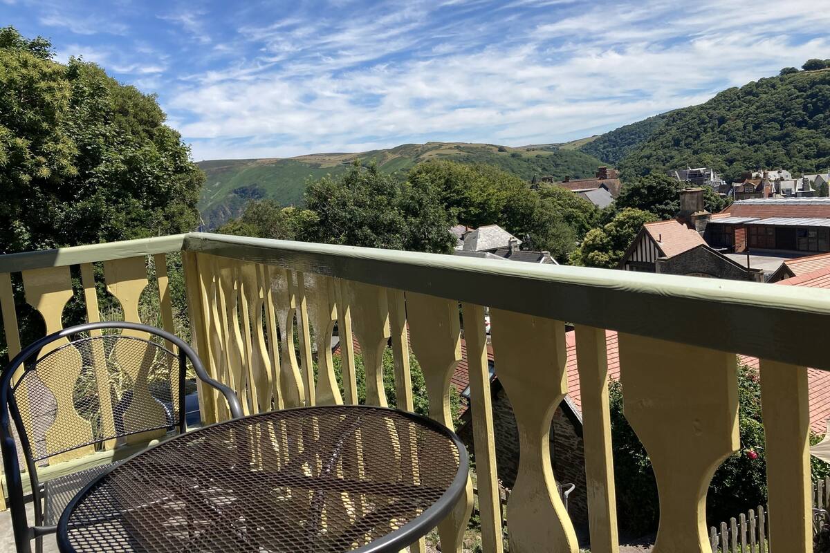 A balcony with a metal chair and a table, overlooking a town with houses and green hills.