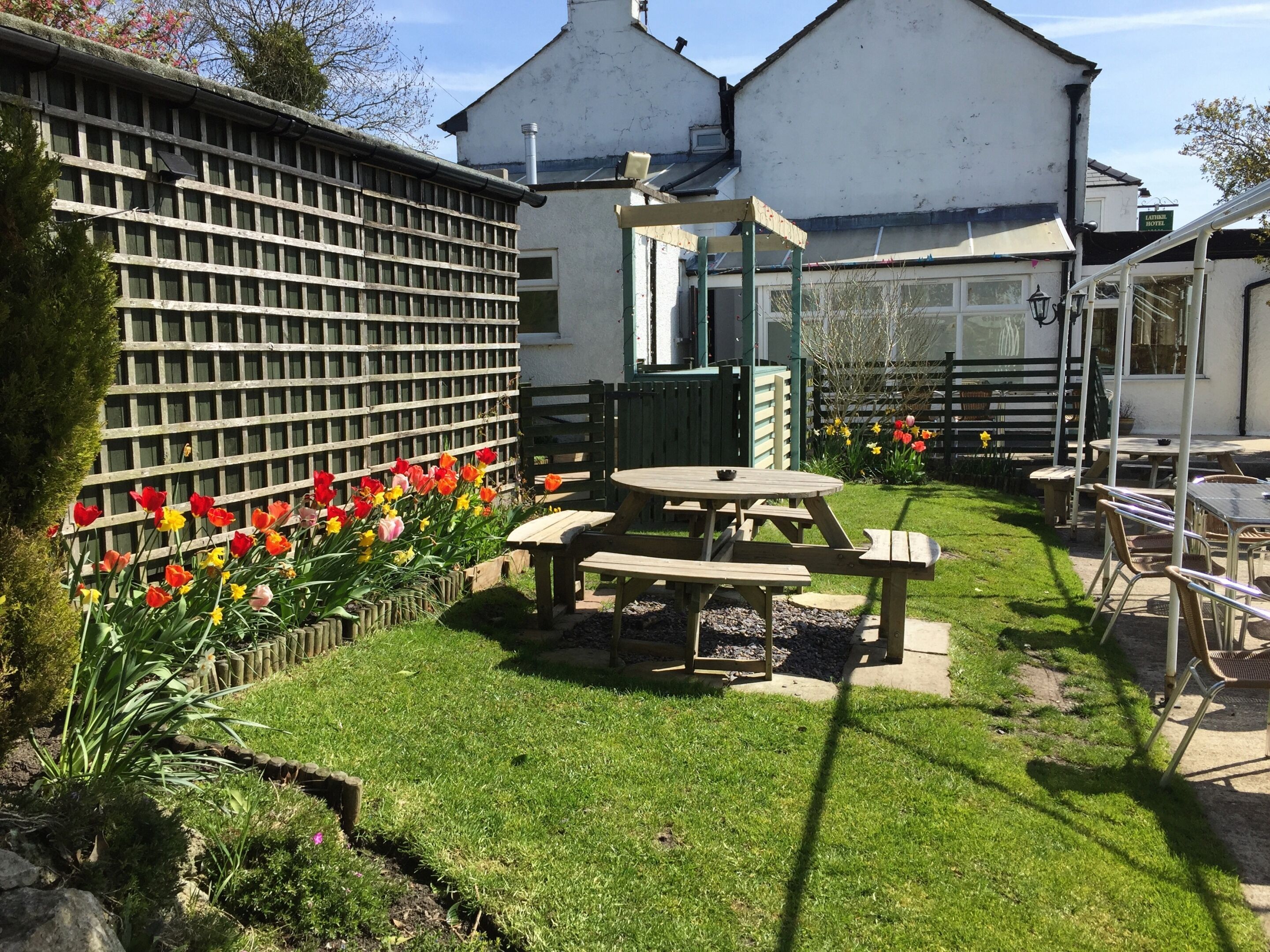 A garden area with a wooden fence, a wooden table and benches, and a variety of flowers.