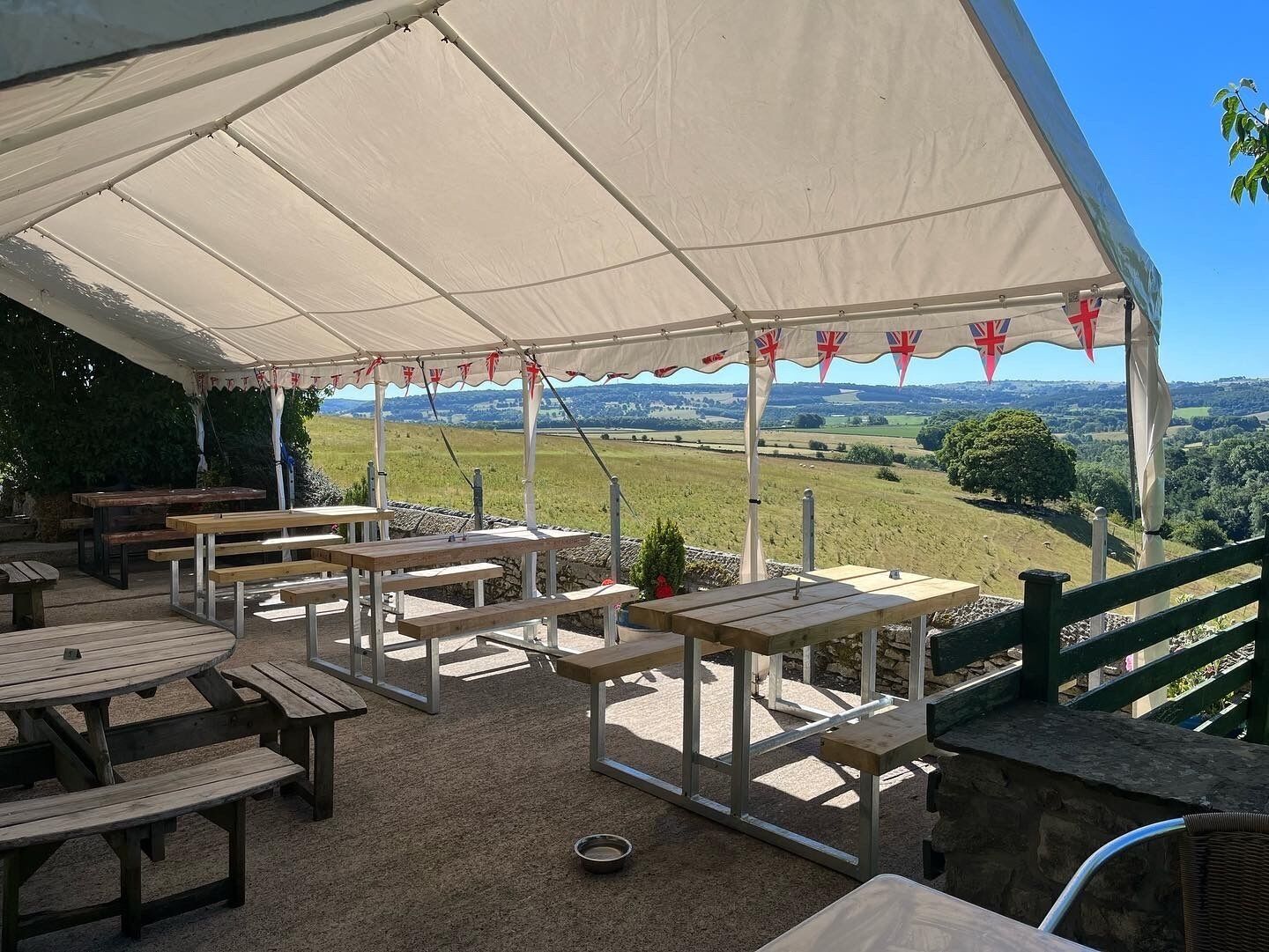 A covered outdoor seating area with picnic tables, a stone wall, and a view of a green landscape.