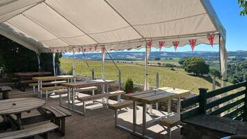 A covered outdoor seating area with picnic tables, a stone wall, and a view of a green landscape.
