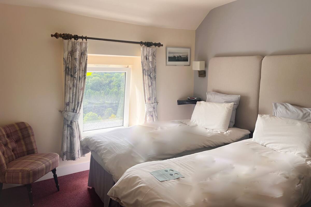 A hotel room with two beds, a chair, a nightstand with a lamp, and a window with curtains.