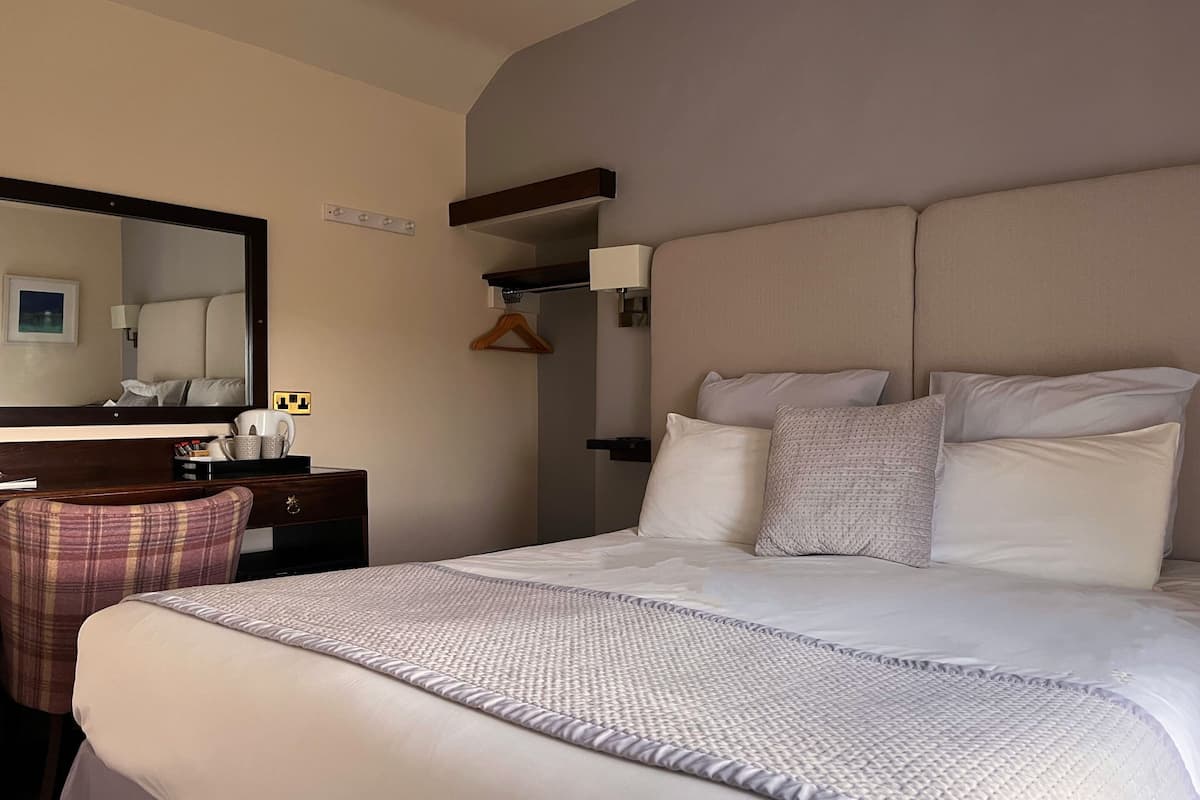 A hotel room with a large bed, a bedside table with a lamp, a mirror, and a small desk with a chair.