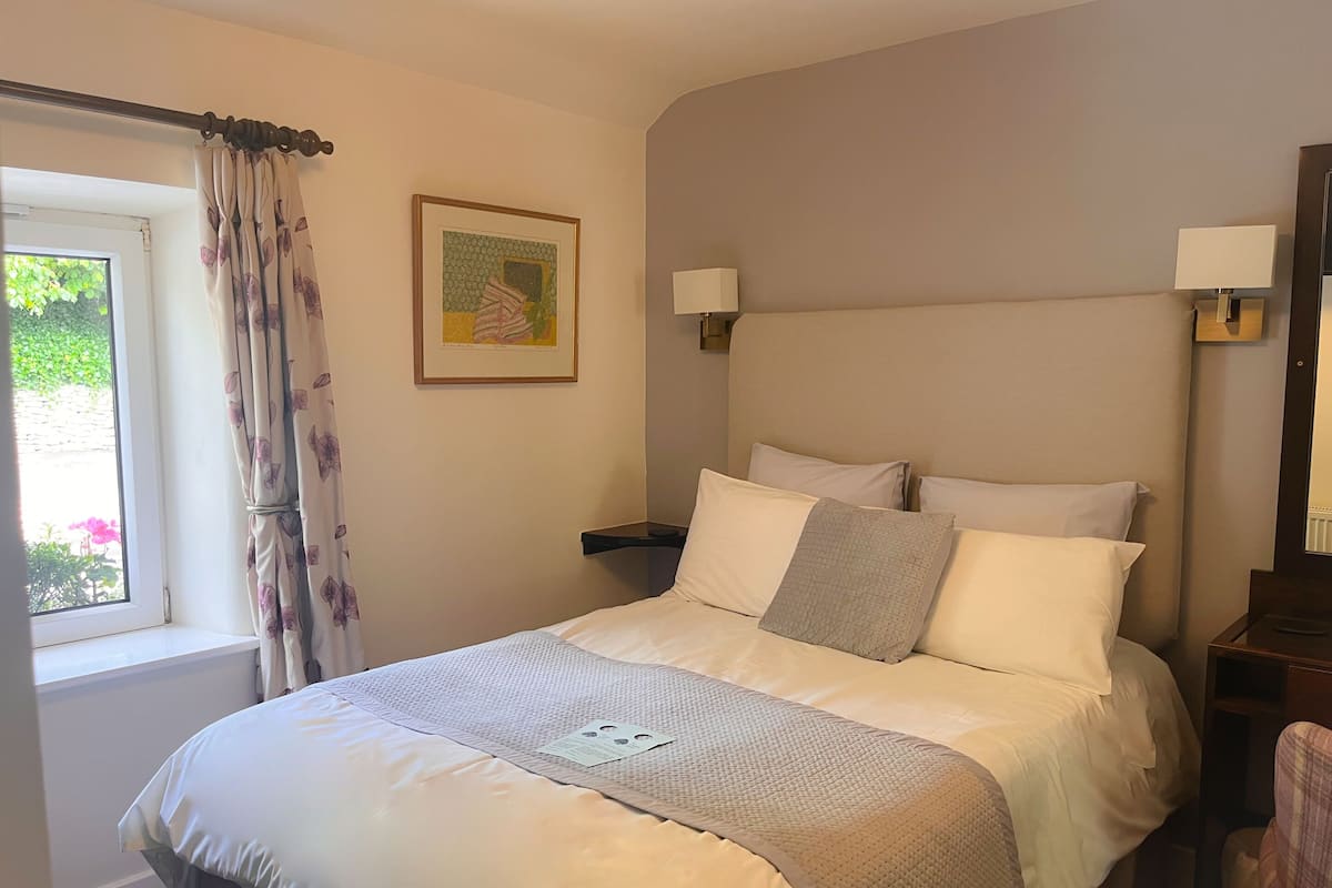 A neatly made bed with a white comforter and pillows, a bedside table with a lamp, a framed picture on the wall, and a window with curtains.