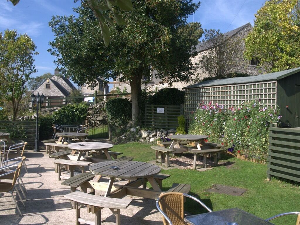 An outdoor patio area with multiple wooden tables and chairs, surrounded by greenery and a stone building in the background.