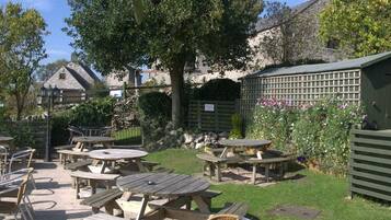 An outdoor patio area with multiple wooden tables and chairs, surrounded by greenery and a stone building in the background.