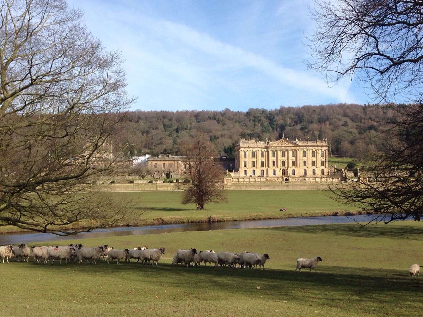 A large, classical building with symmetrical architecture, a grassy field, and a river in the foreground.