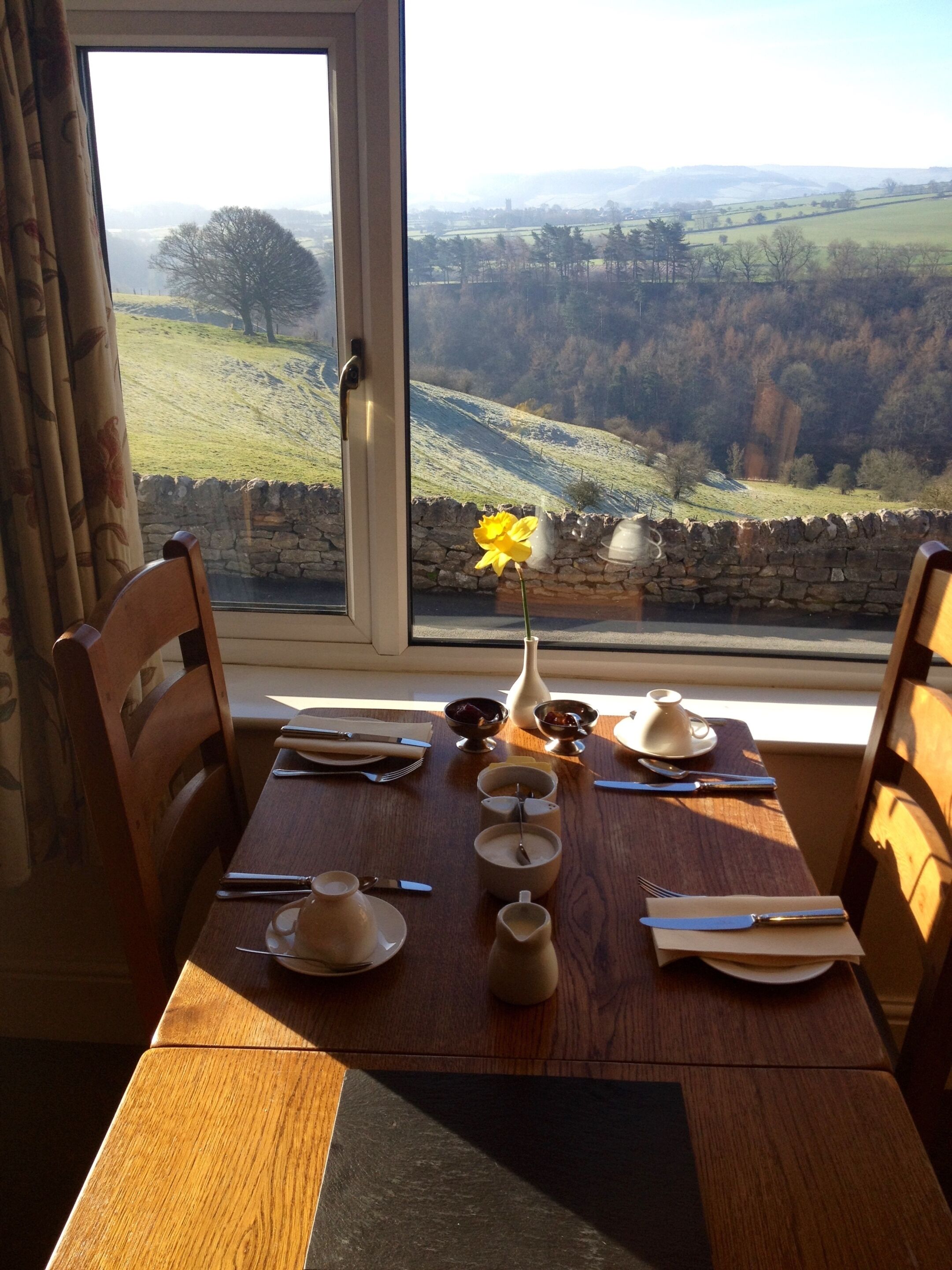 A dining table set for two with a view of a rural landscape through a window.