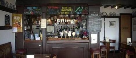 A bar area with wooden beams, a counter displaying various bottles, and a menu board.