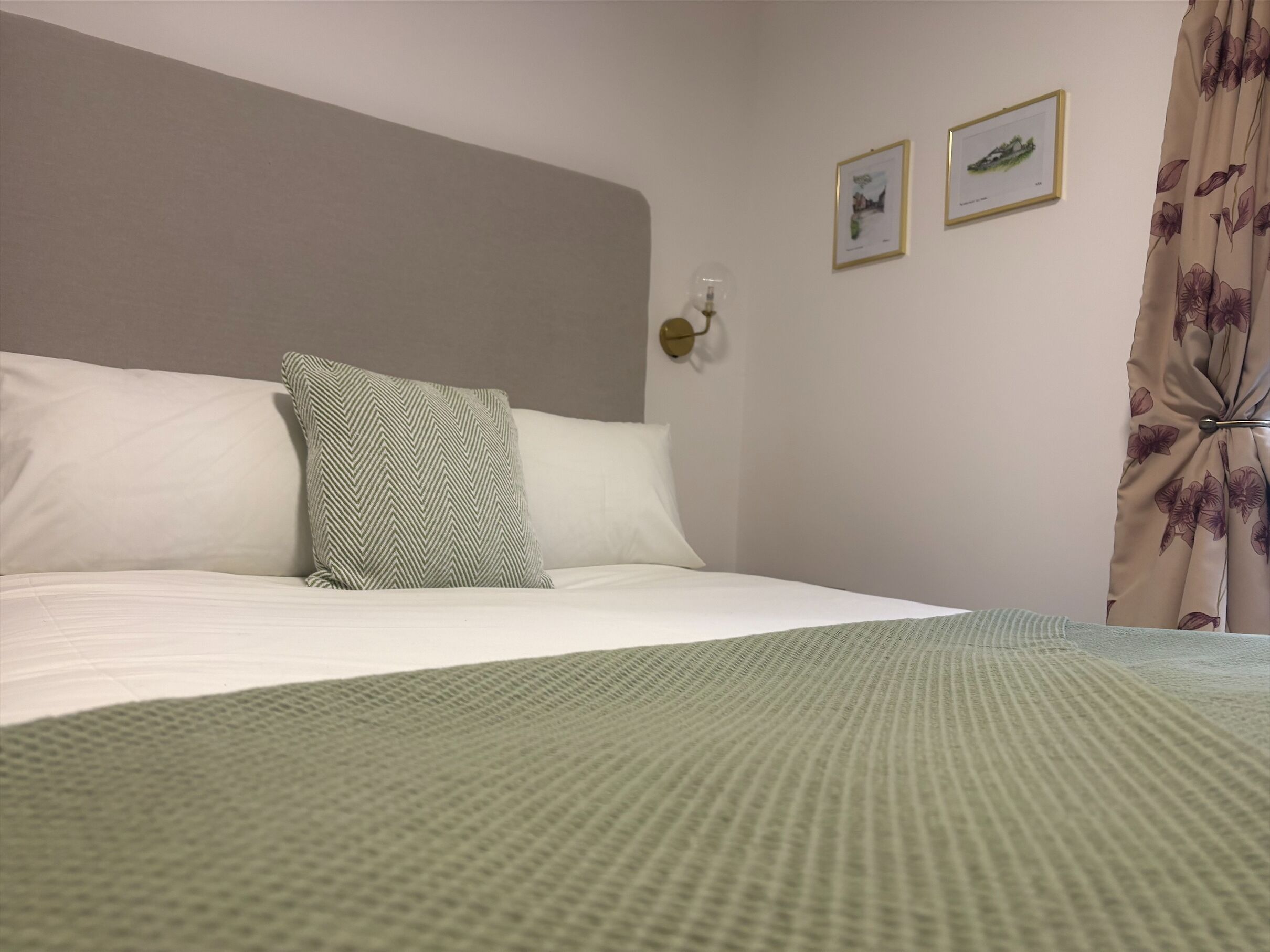 Un lit bien fait, recouvert d’une couverture verte à texture, avec des oreillers blancs et un coussin à motifs. Deux tableaux encadrés sont accrochés au mur, et un luminaire est fixé au plafond.