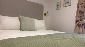 Un lit bien fait, recouvert d’une couverture verte à texture, avec des oreillers blancs et un coussin à motifs. Deux tableaux encadrés sont accrochés au mur, et un luminaire est fixé au plafond.