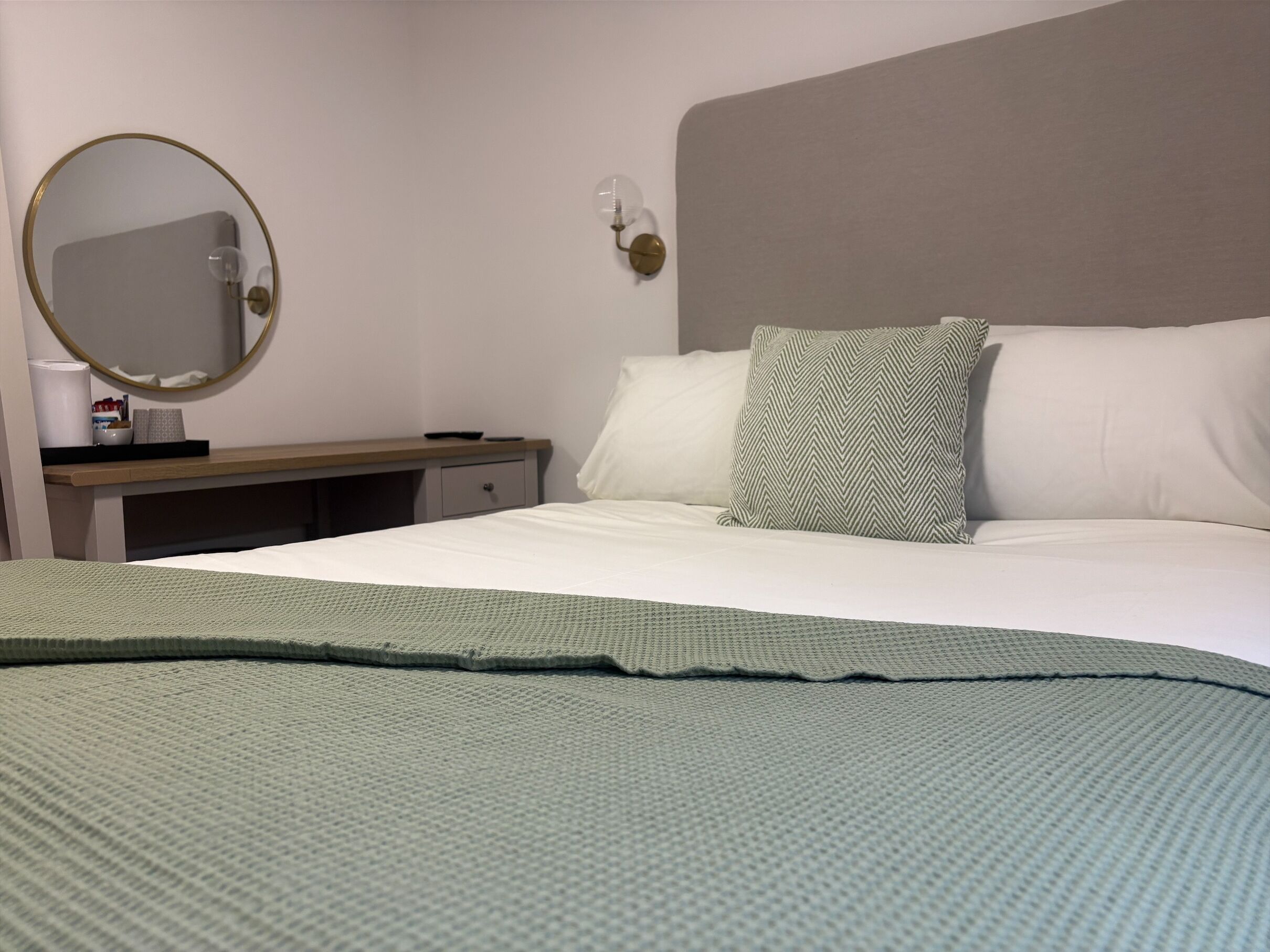 Una habitación de hotel con una cama, una mesita de noche, un espejo y una lámpara fijada a la pared.