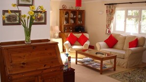 A living room with a wooden cabinet, a glass coffee table, a beige sofa with red cushions, a white armchair, and a floral rug. - Leworthy Farmhouse (Holsworthy)