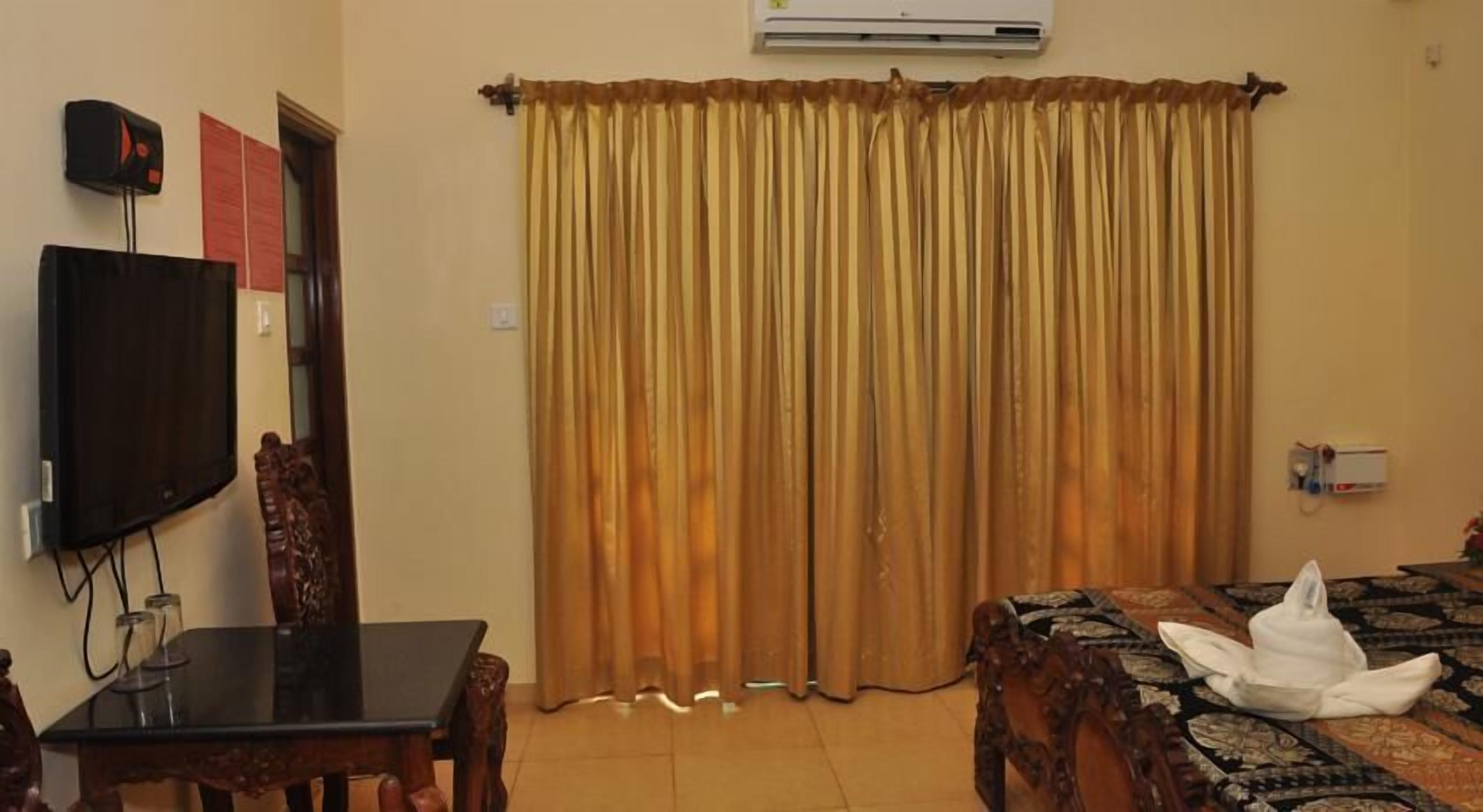 A room with a television, a table, a chair, a bed with a patterned bedspread, and a curtain.
