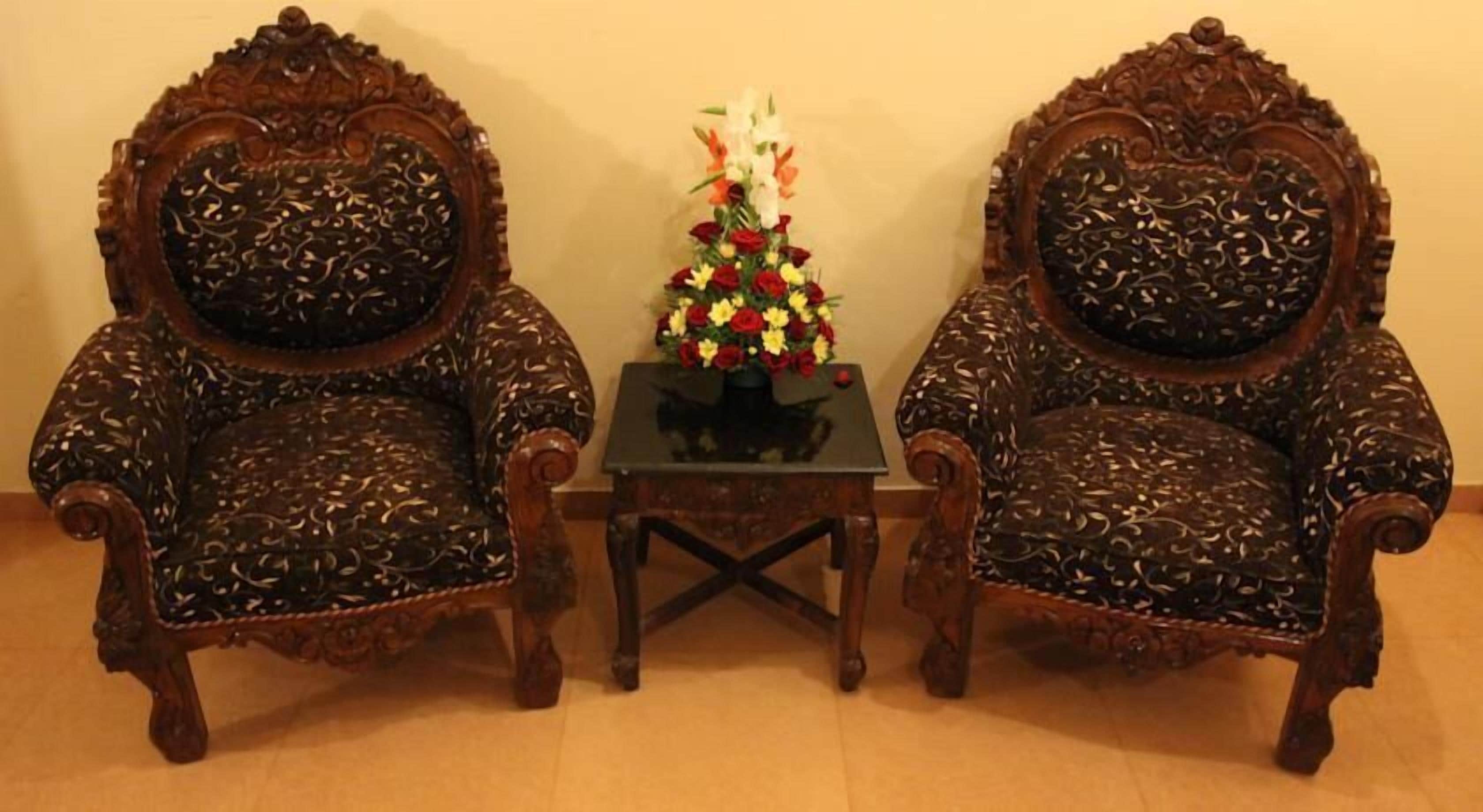 Two ornate wooden chairs with intricate carvings, a small table with a floral arrangement, and a plain wall in the background.