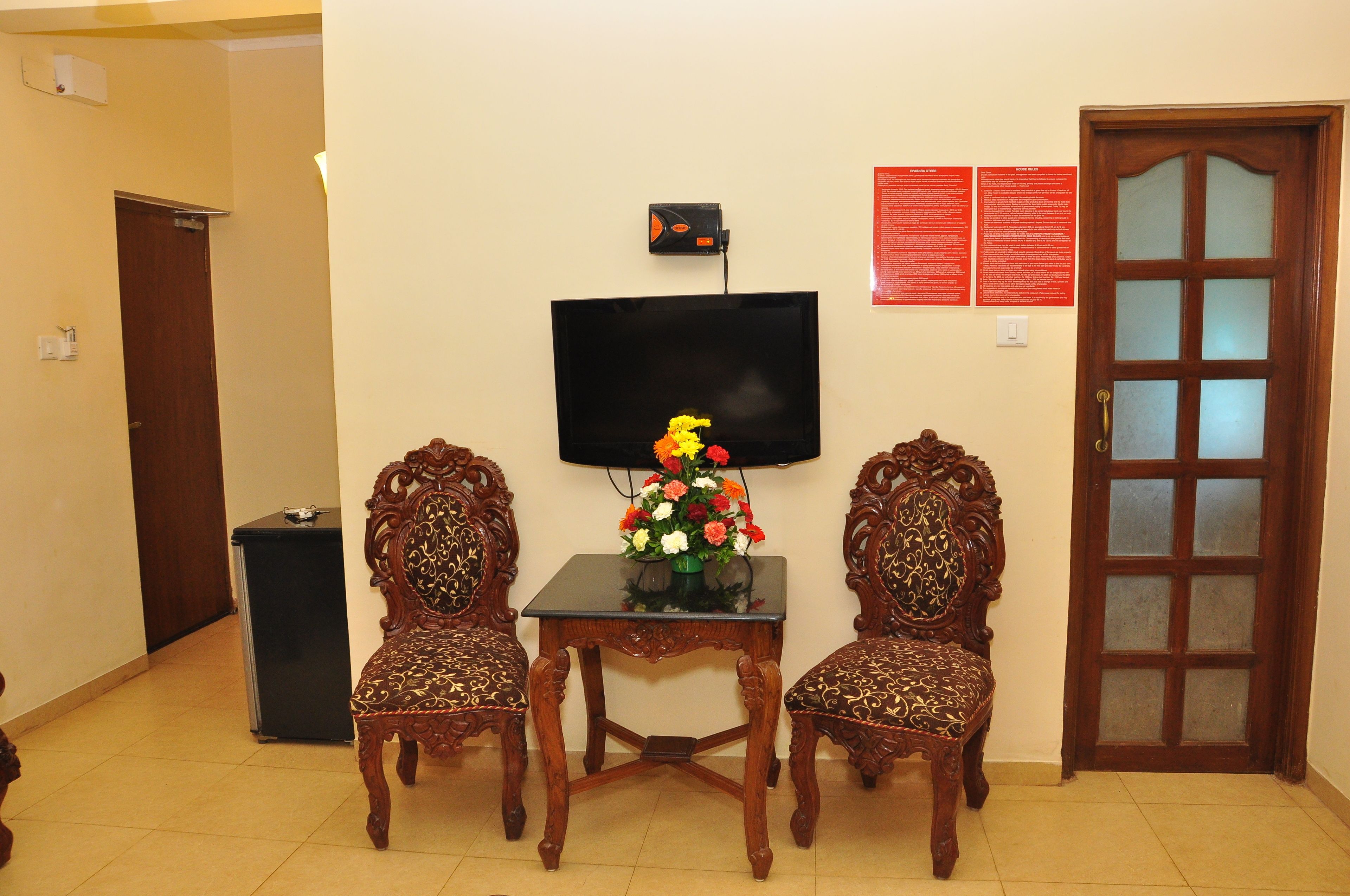 A room with a television on the wall, a small table with a vase of flowers, two ornate chairs, and a door with a glass panel.