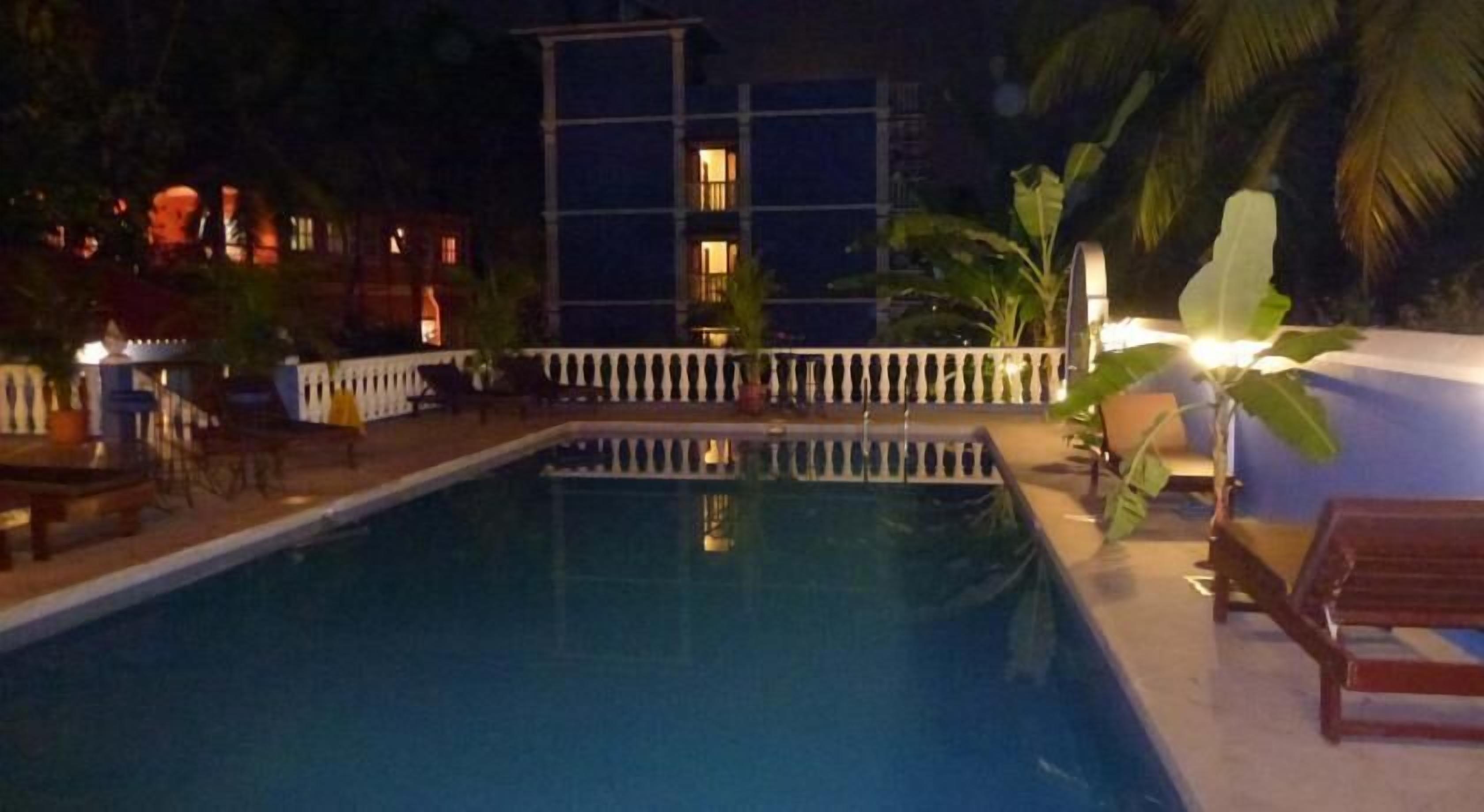 A swimming pool with a white fence, lounge chairs, and a building with illuminated windows in the background.